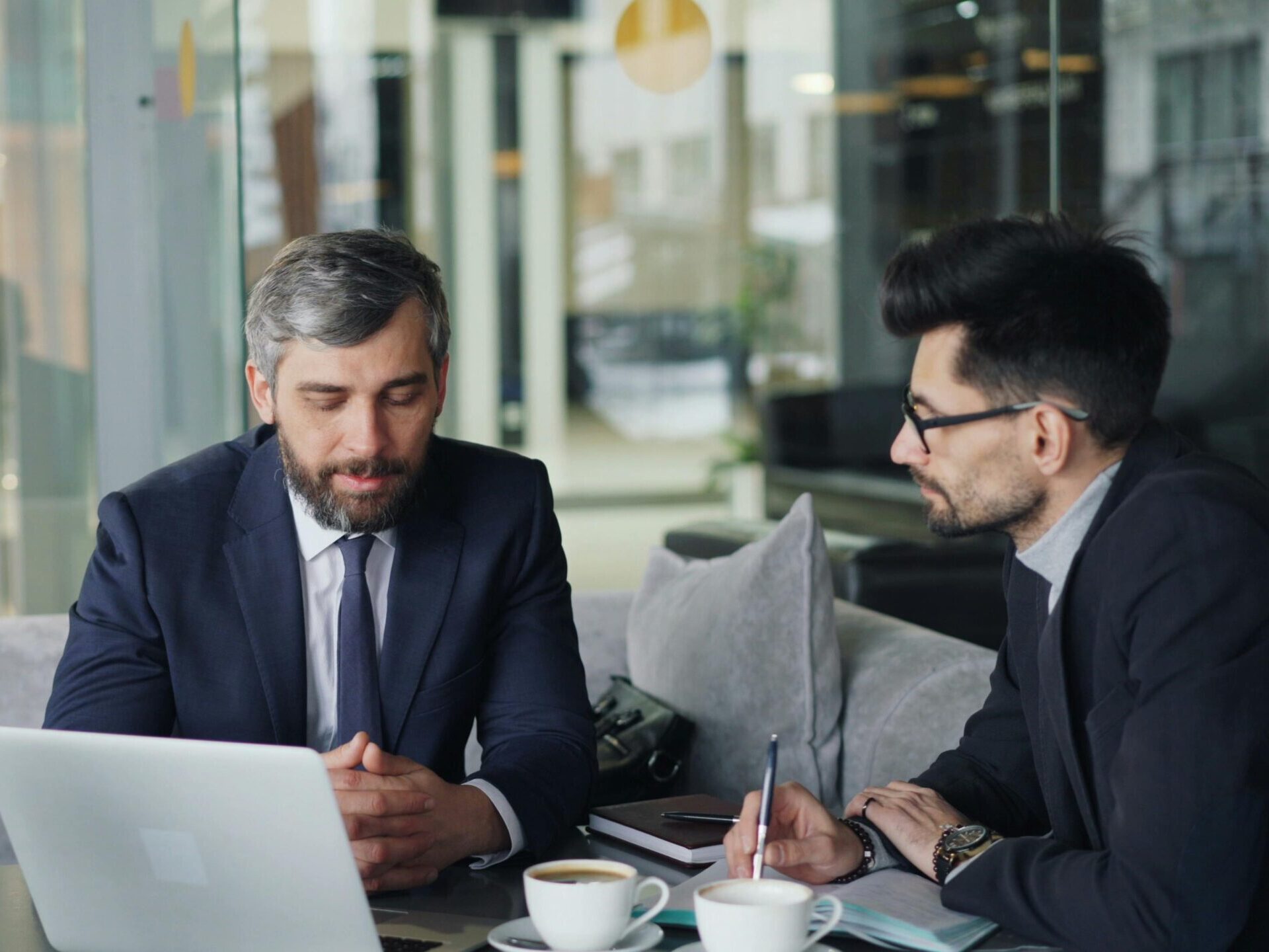 Two businessmen in suits discussing work at a cafe with laptops and coffee.