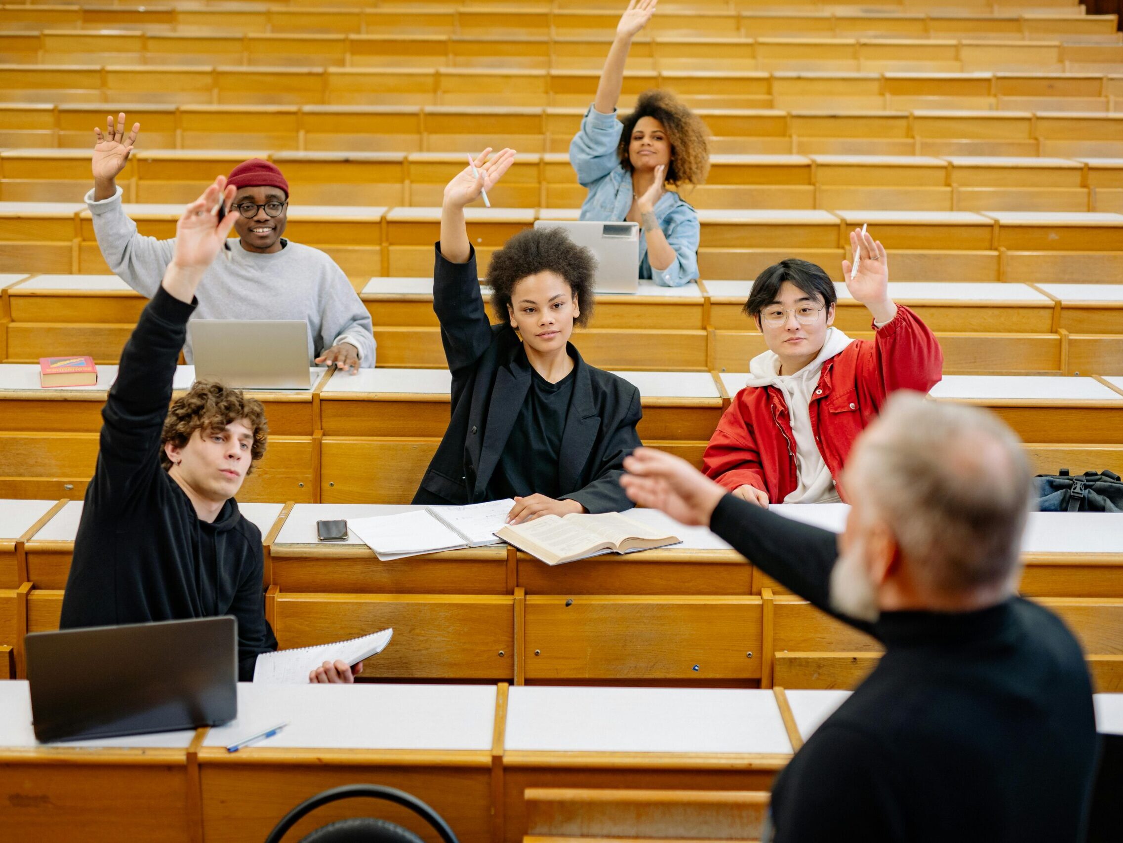 A diverse group of university students participating in a lecture, raising hands to engage with the professor.