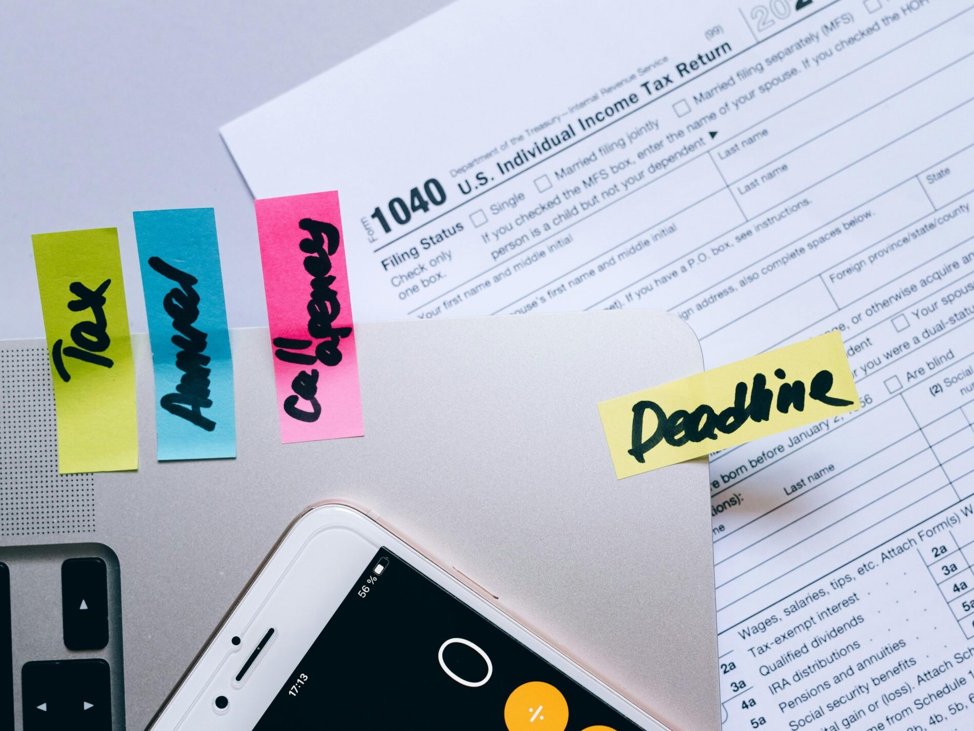 Office desk flat lay showing tax documents, calculator app on smartphone, sticky notes, and paperclips.
